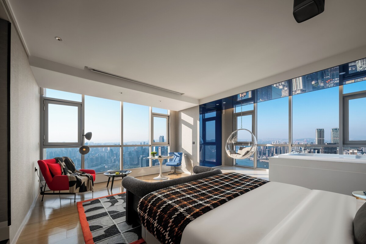 A modern hotel room with a large bed, a red chair, a desk, and a view of a cityscape through large windows.