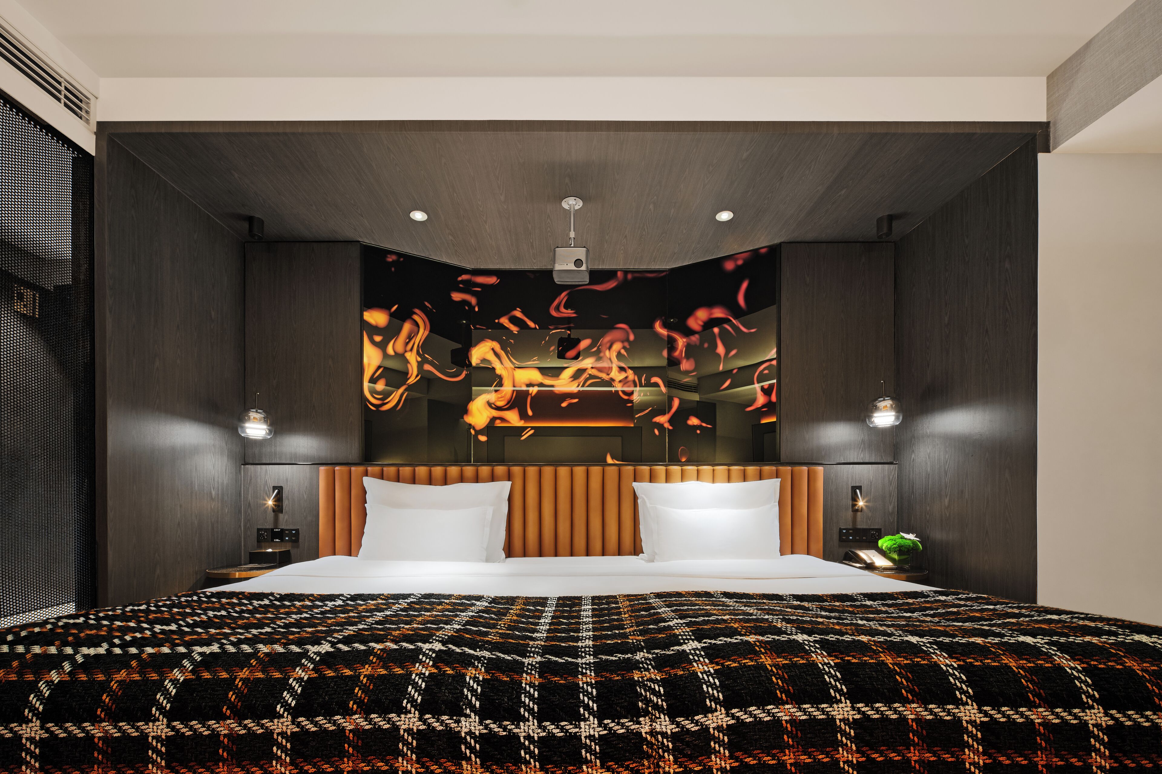 A modern hotel room with a large bed, a headboard with vertical slats, recessed ceiling lights, and a wall-mounted projector.