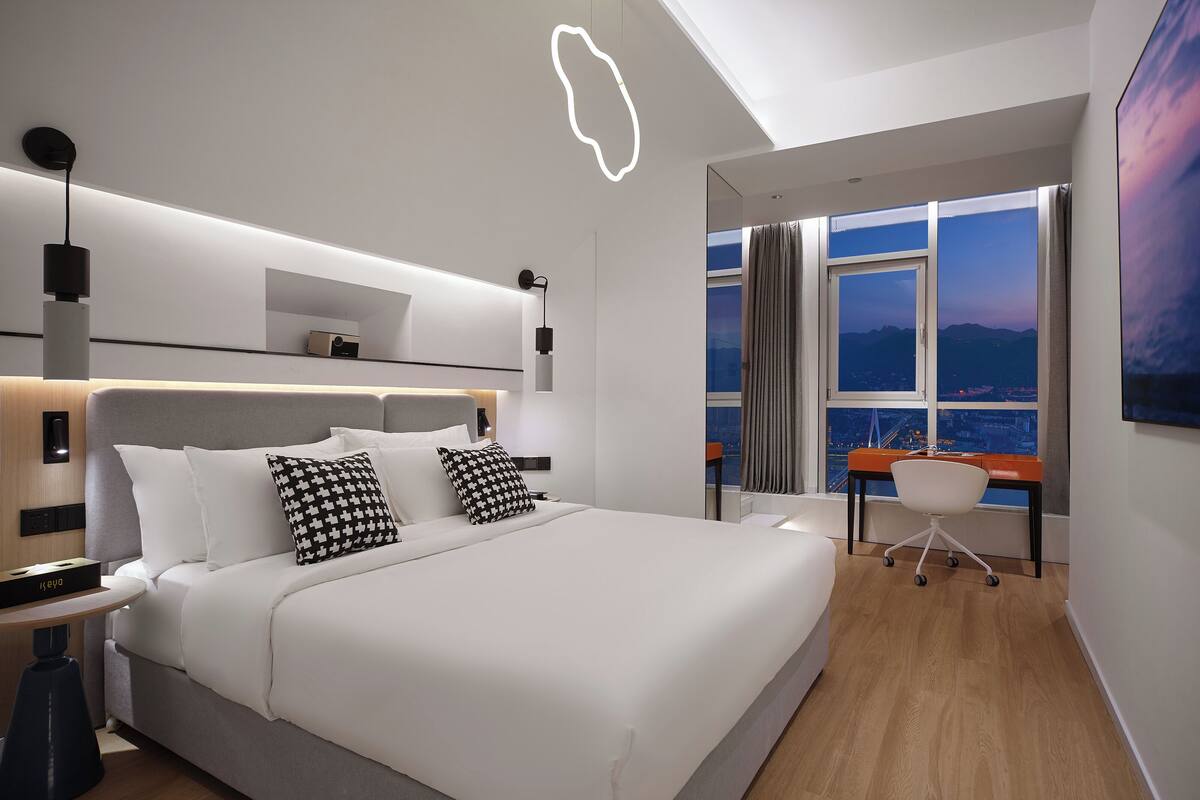 Une chambre d’hôtel moderne dotée d’un grand lit, de tables de chevet, d’un bureau avec une chaise et offrant une vue sur le paysage urbain grâce à de grandes fenêtres.