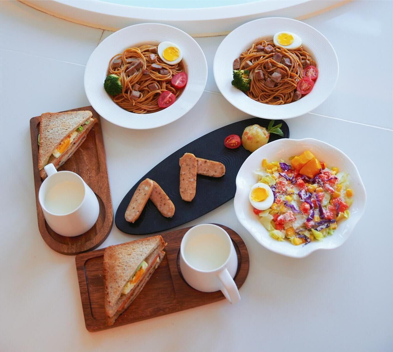 Un plato de fideos, huevos y verduras, acompañado de sándwiches y un vaso de leche.