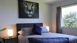 A bedroom with a bed, a nightstand with a lamp, a painting of a lion's face, and a window with curtains. - Safe Haven Hillside Retreat (Maungatapere)