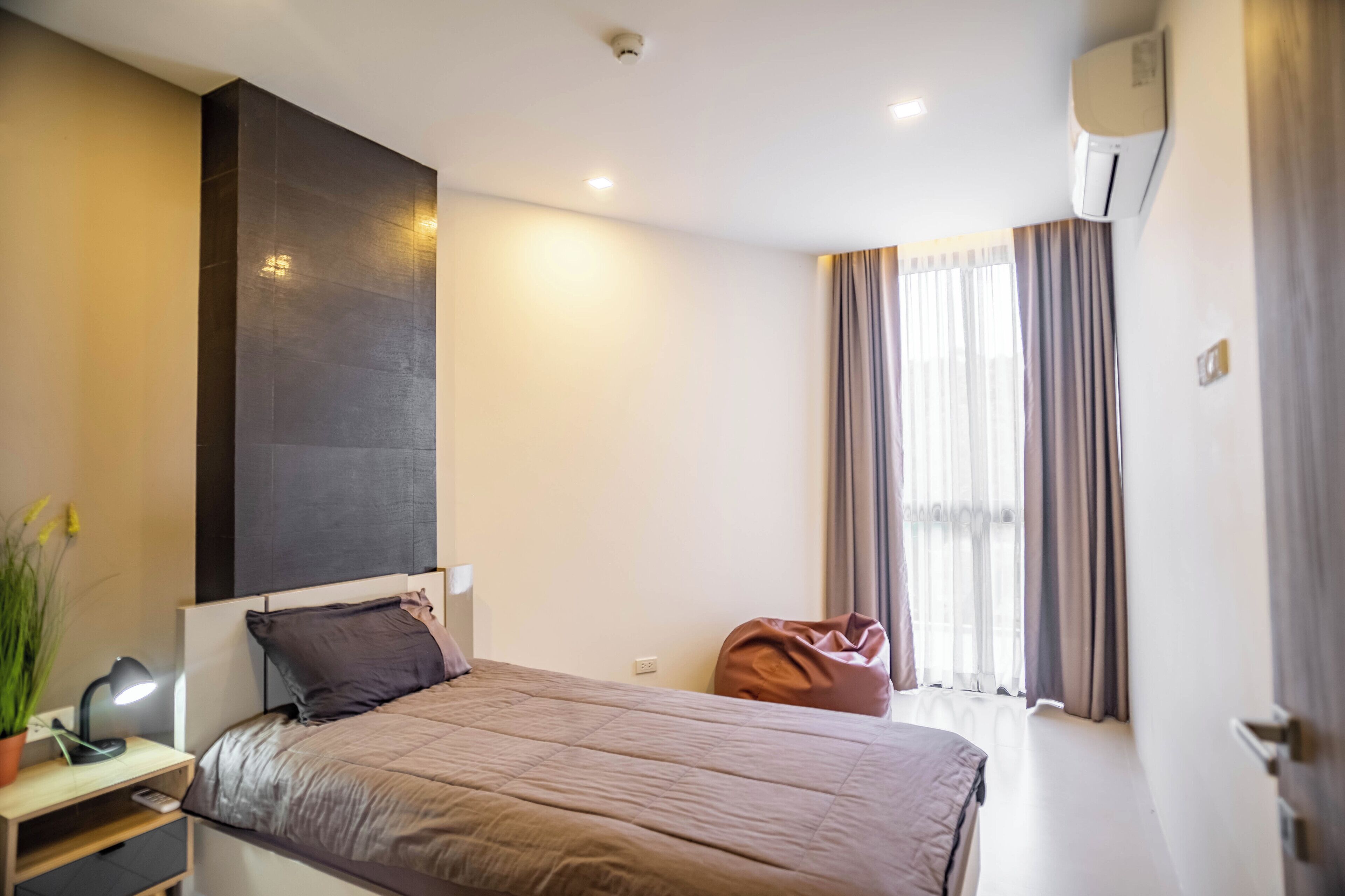 A modern hotel room with a bed, bedside table, lamp, air conditioning unit, and a window with curtains.
