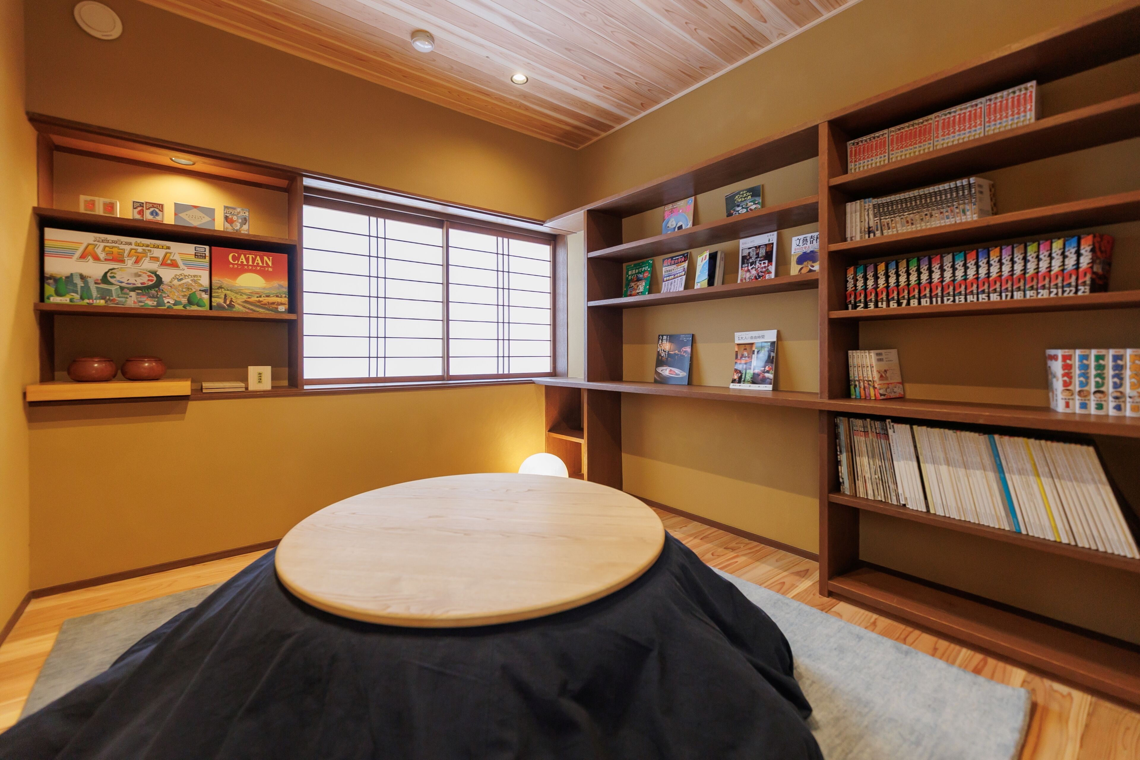 A room with a tatami mat, wooden shelves with books and games, a window with blinds, and a ceiling with recessed lighting.