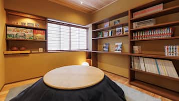 A room with a tatami mat, wooden shelves with books and games, a window with blinds, and a ceiling with recessed lighting.