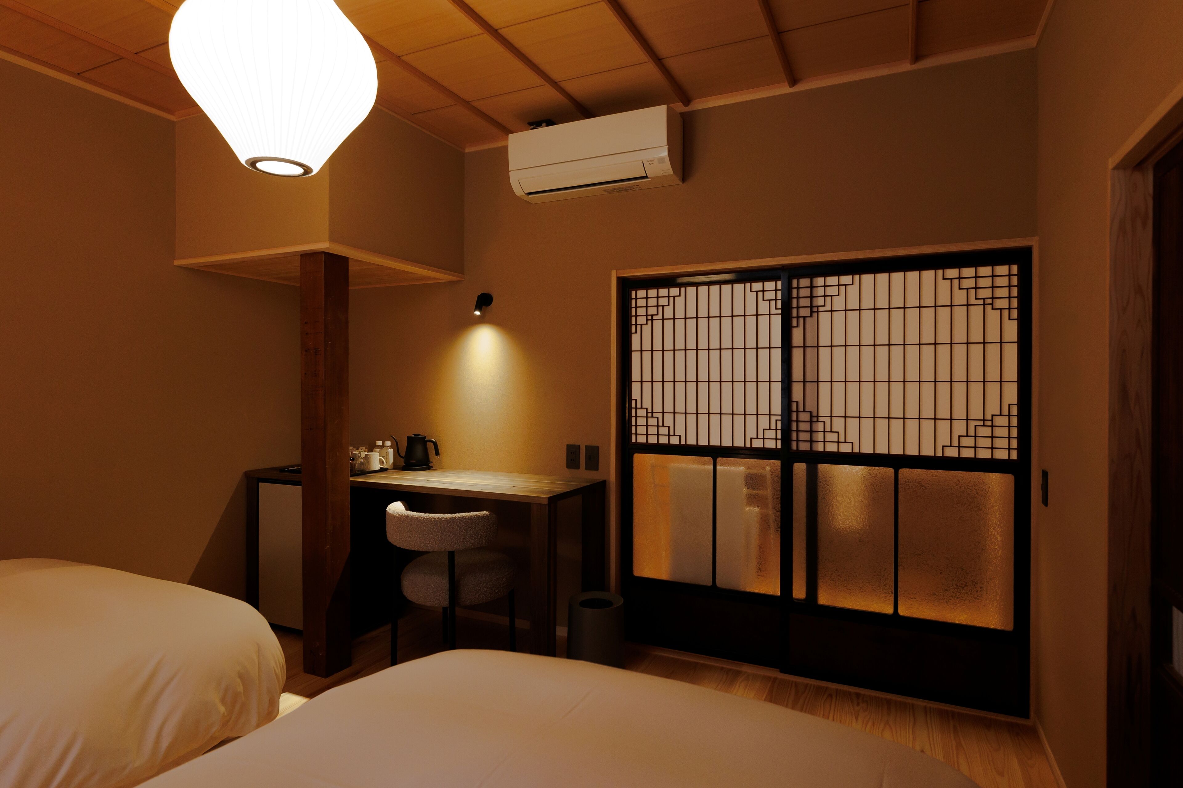 Une chambre d’hôtel équipée d’un lit, d’un bureau, d’une chaise, d’un climatiseur fixé au mur et d’une fenêtre de style traditionnel avec des croisillons.