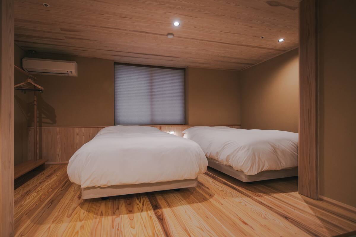 Ein Schlafzimmer mit zwei Betten, Holzboden, einem Fenster mit Jalousien und einer Klimaanlage.