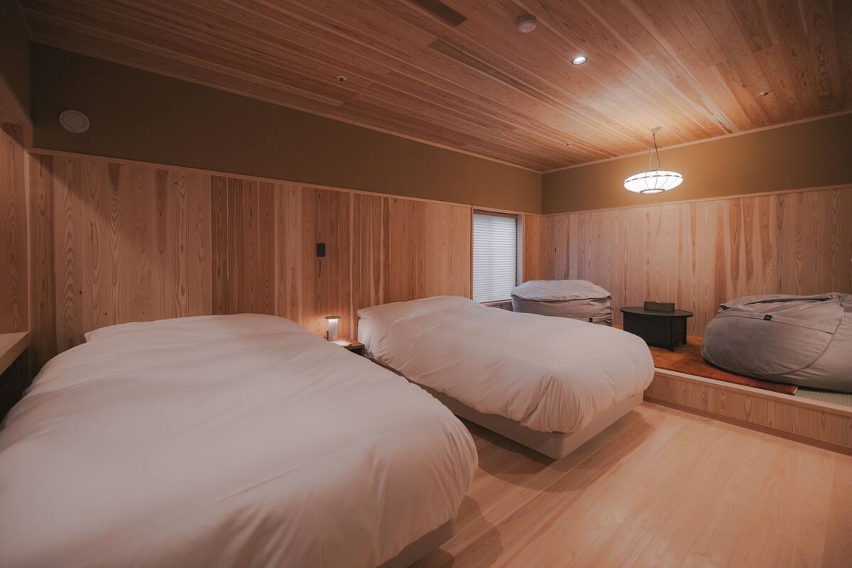 Ein Zimmer mit zwei Betten, Holzwandverkleidung, einem Fenster mit Jalousien, einem kleinen Tisch und einem Sitzsack.
