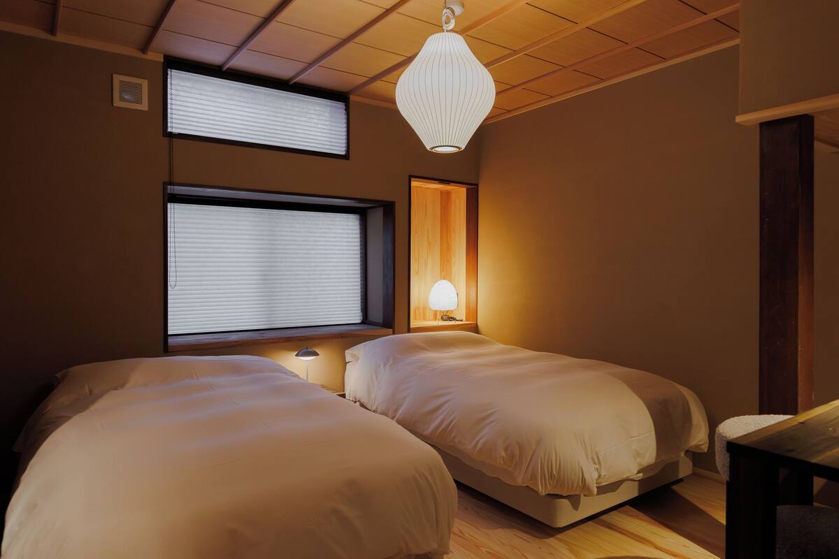 Ein Zimmer mit zwei Betten, einem Fenster mit Jalousien, einer Wandlampe und einer Deckenleuchte.