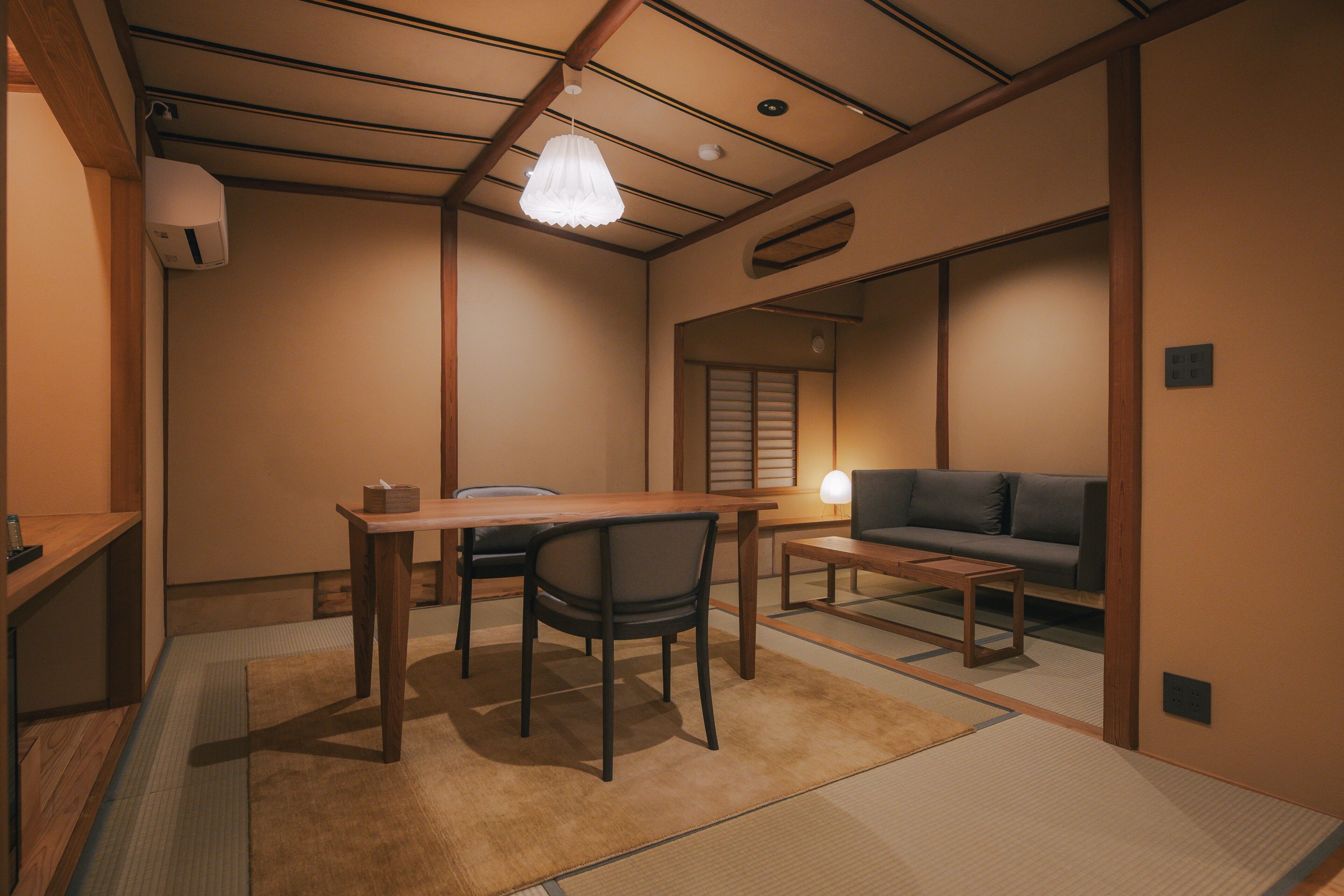 A traditional Japanese room with tatami flooring, a low table, chairs, a sofa, and a hanging light fixture.