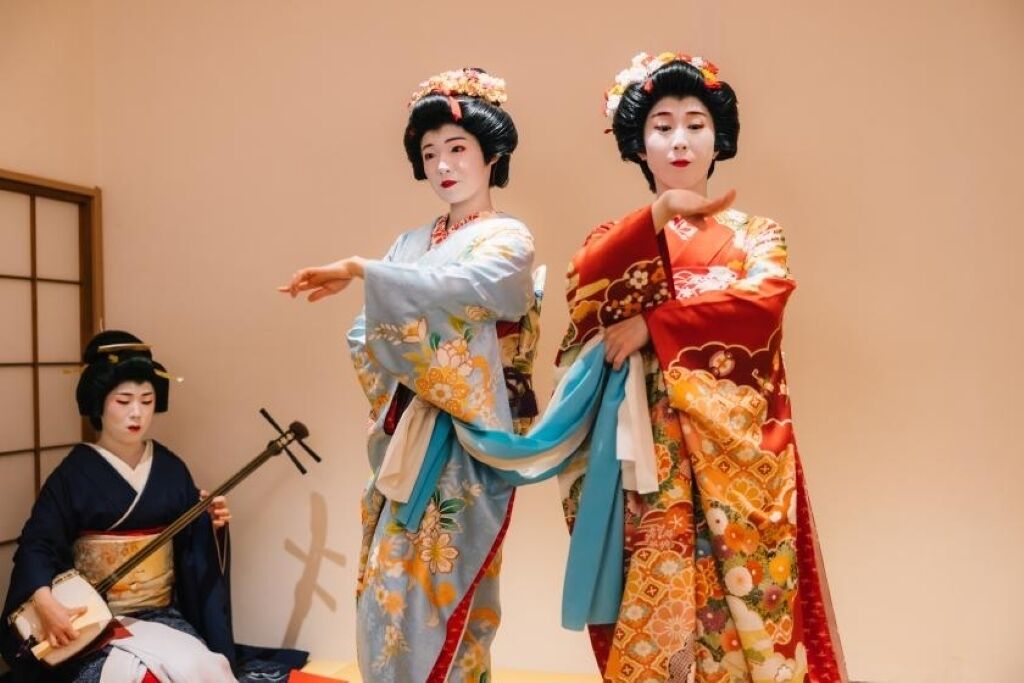 Two individuals in traditional attire, one playing a stringed instrument, in a room with a simple, unadorned wall and a visible window.