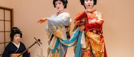 Two individuals in traditional attire, one playing a stringed instrument, in a room with a simple, unadorned wall and a visible window.