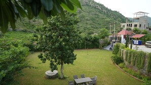 A grassy outdoor area with a table and chairs, a tree, and a building with a red roof in the background. - Cozy Cliff By Marwar Royal – Private Villa with Pool (Barodiya)