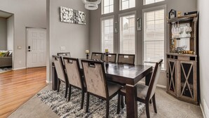 A dining area with a large wooden table, six chairs, and a chandelier. There are large windows with blinds, a door, and a cabinet with glassware. - 6 Mi to Buckley Space Force Base: Getaway w/ Patio (Aurora)