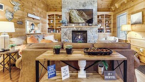 Ein gemütliches Blockhaus-Interieur mit Kamin aus Stein, einem darüber montierten Fernseher und einer einladenden Sitzecke mit Sofa und Couchtisch.