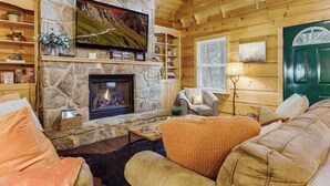 Ein gemütliches Blockhaus-Interieur mit Kamin aus Stein, Holzwänden, einer komfortablen Sitzecke und einem Fernseher über dem Kamin.