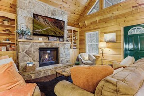A cozy log cabin interior with a stone fireplace, wooden walls, a comfortable seating area, and a television mounted above the fireplace. - Waterfall Log Cabin: Private Hot Tub & Game Room (Gatlinburg)