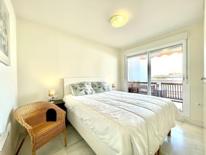 A bedroom with a bed, a wicker chair, a nightstand with a lamp, and a sliding glass door leading to an outdoor balcony. - Belvilla by OYO Agua Serena IV (Roquetas de Mar)