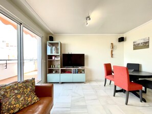 A living room with a TV, a bookshelf, a dining area with chairs, and a large window. - Belvilla by OYO Agua Serena IV (Roquetas de Mar)