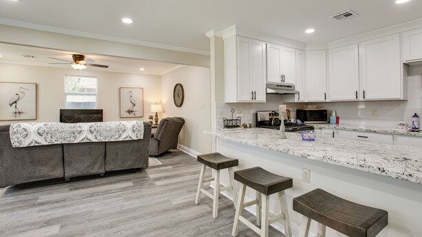 A modern kitchen with white cabinets, a granite countertop, and a built-in stove. There are two bar stools and a dining area with a gray sofa and a table.