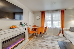 Ein modernes Wohnzimmer mit Kamin, einem Essbereich mit orangefarbenen Stühlen, einem weißen Sofa und einem großen Flachbildfernseher an der Wand.