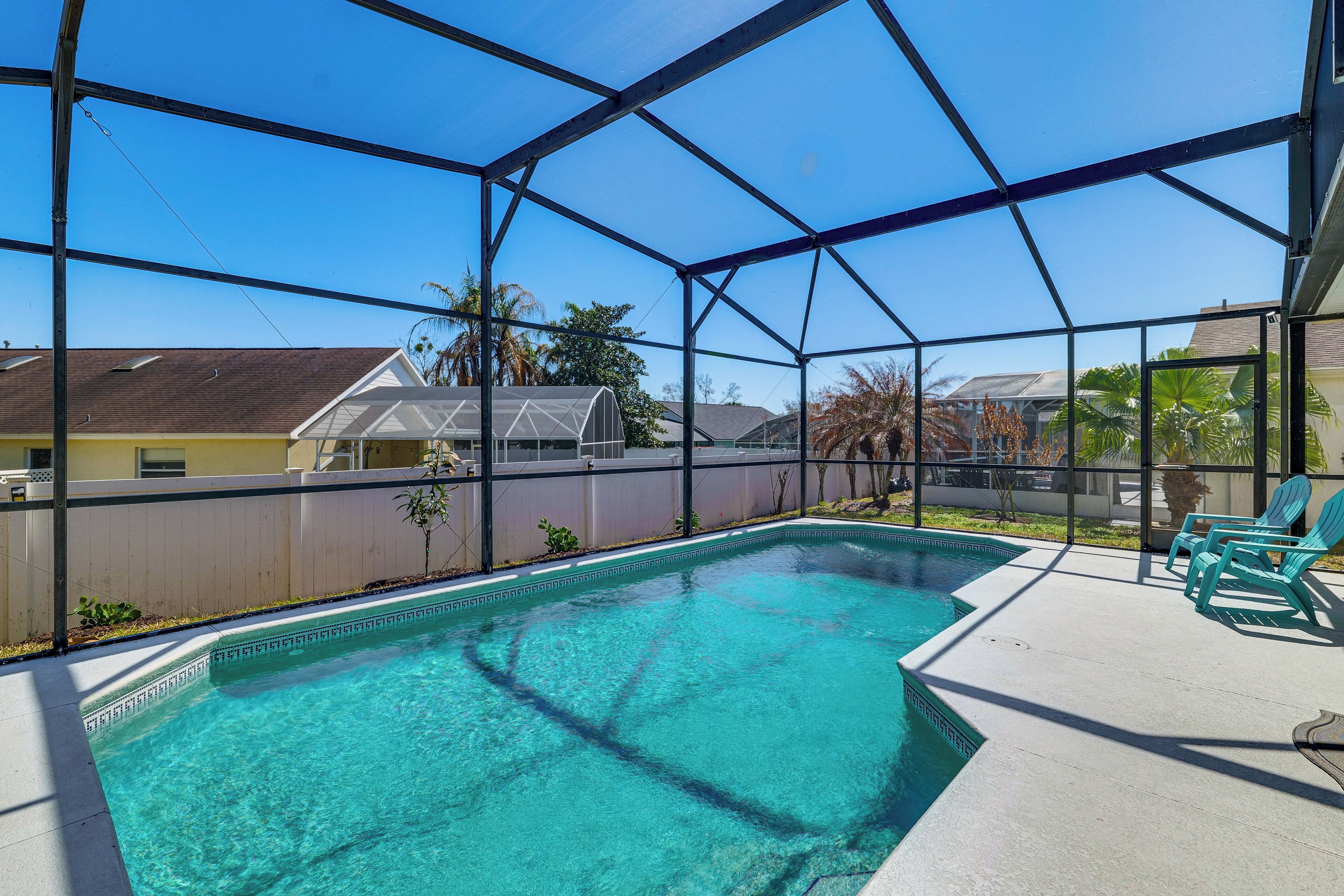 Une piscine d’un bleu éclatant, protégée par une paroi de verre, entourée d’une terrasse aménagée avec des chaises et offrant une vue sur les maisons voisines.