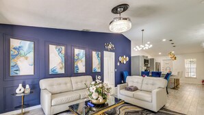 A modern living room with two white sofas, a glass coffee table, and a blue accent wall with framed artwork. - 5 Mi to Disney: Renovated Group Getaway, Game Room (Kissimmee)