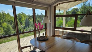 Un comedor con mesa y sillas de madera, ventanas amplias con vistas a zonas verdes y una lámpara colgante.