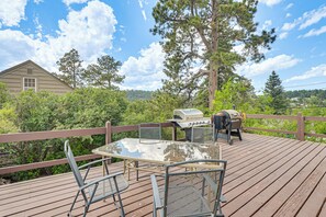 Eine Holzterrasse mit Glastisch und -stühlen, einem Grill und Blick auf ein Haus und Bäume.