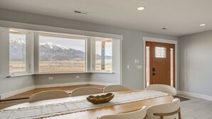 Una sala da pranzo con un tavolo grande, sei sedie, una porta di legno e vista sulle montagne attraverso ampie finestre.