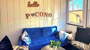 Un sofá azul con una decoración en forma de corazón, una mesa redonda con una planta y una ventana que da a vistas de árboles.