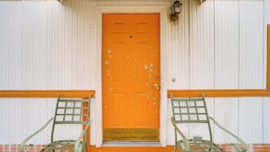Un edificio con una puerta naranja brillante, dos sillas metálicas verdes en la parte delantera y un exterior blanco y naranja.