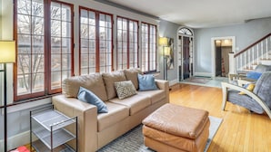 Ein geräumiges Wohnzimmer mit einem beigen Sofa, einem Hocker und einer kleinen Metallregal. Große Fenster mit Holzrahmen sorgen für viel Tageslicht.