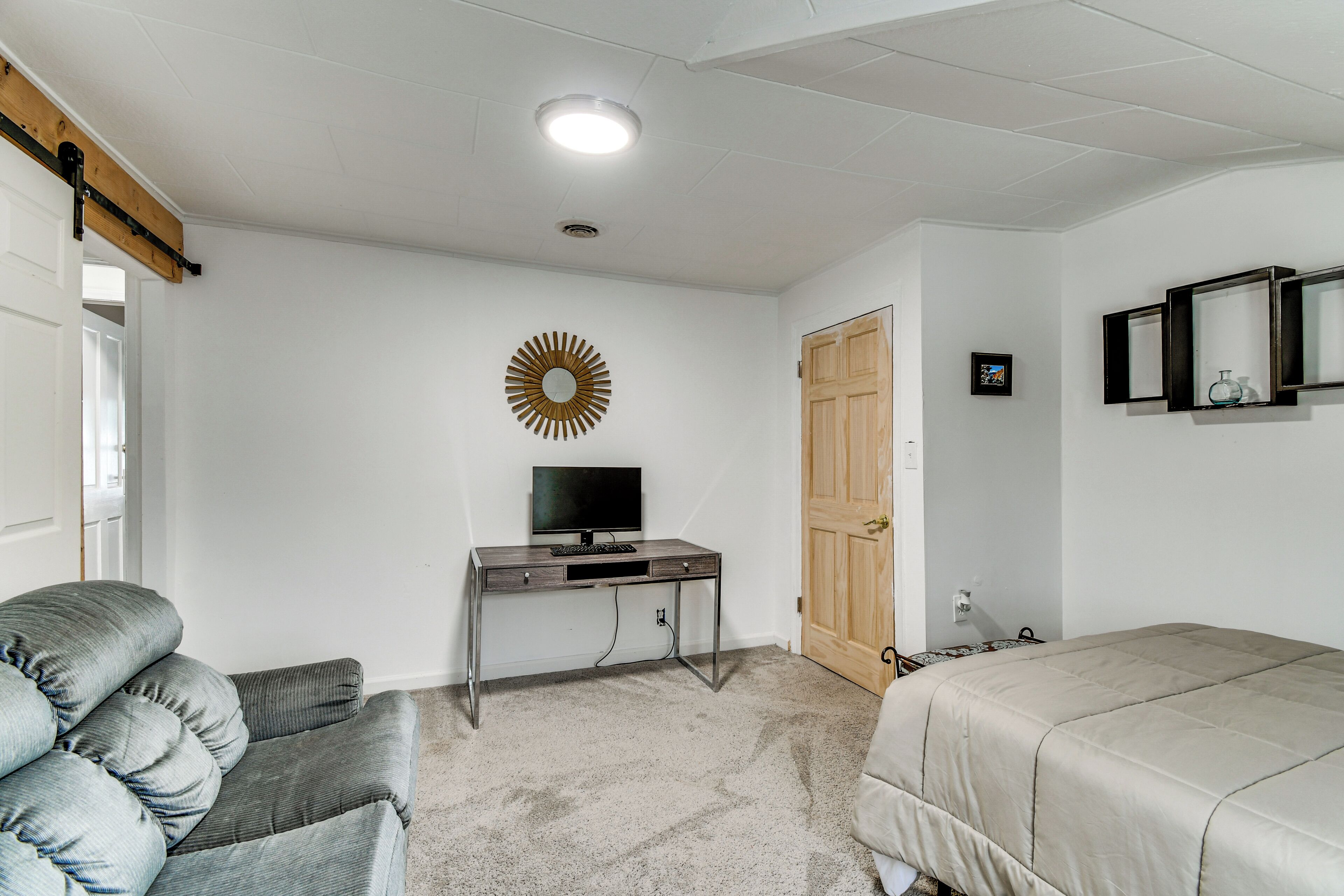 Una habitación con escritorio, silla, cama, puerta y espejo fijado a la pared.