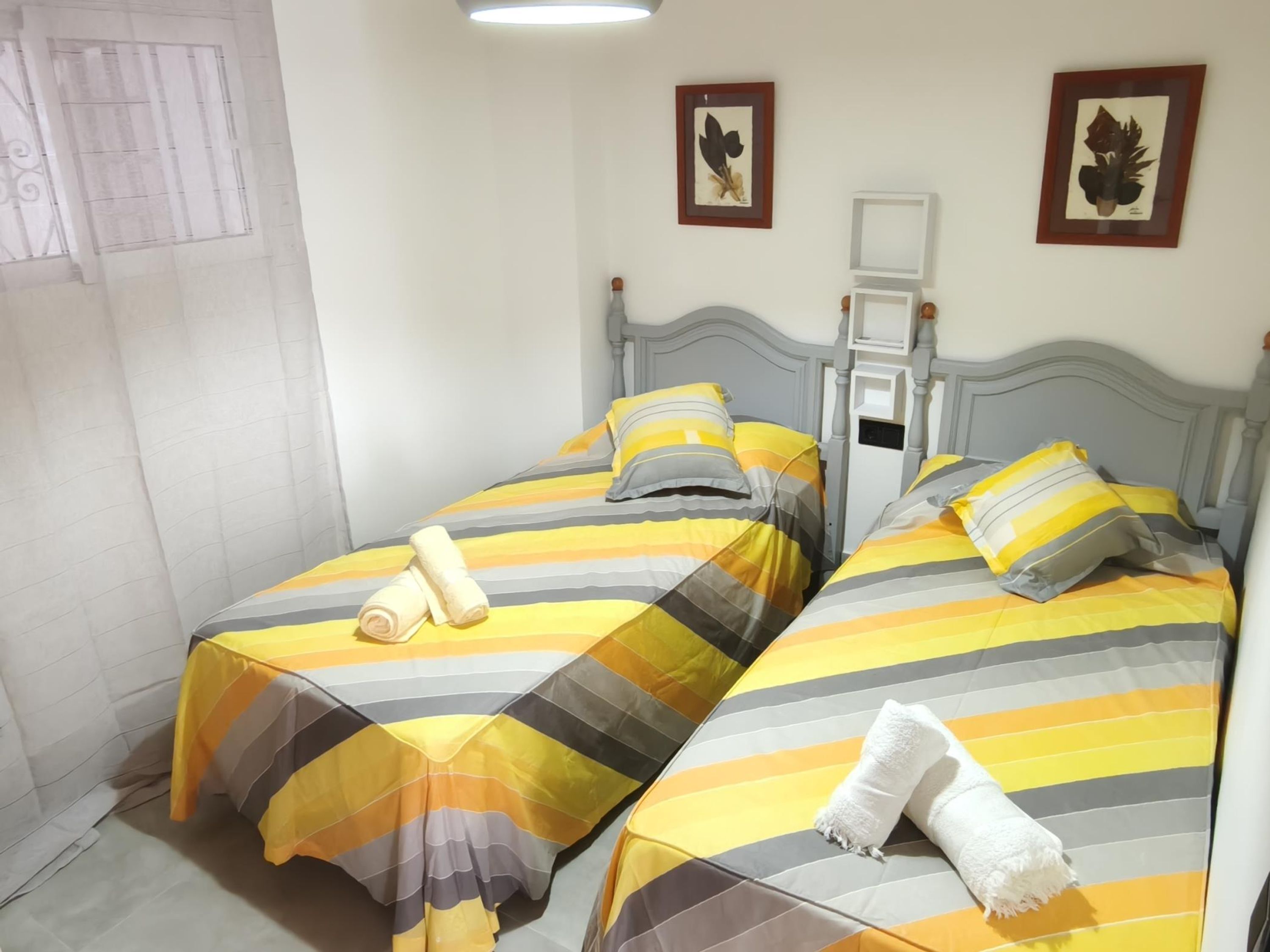 Una habitación con dos camas individuales, cada una con ropa de cama y almohadas a rayas. Hay impresiones botánicas enmarcadas en la pared, un estante blanco pequeño y una ventana con cortina.