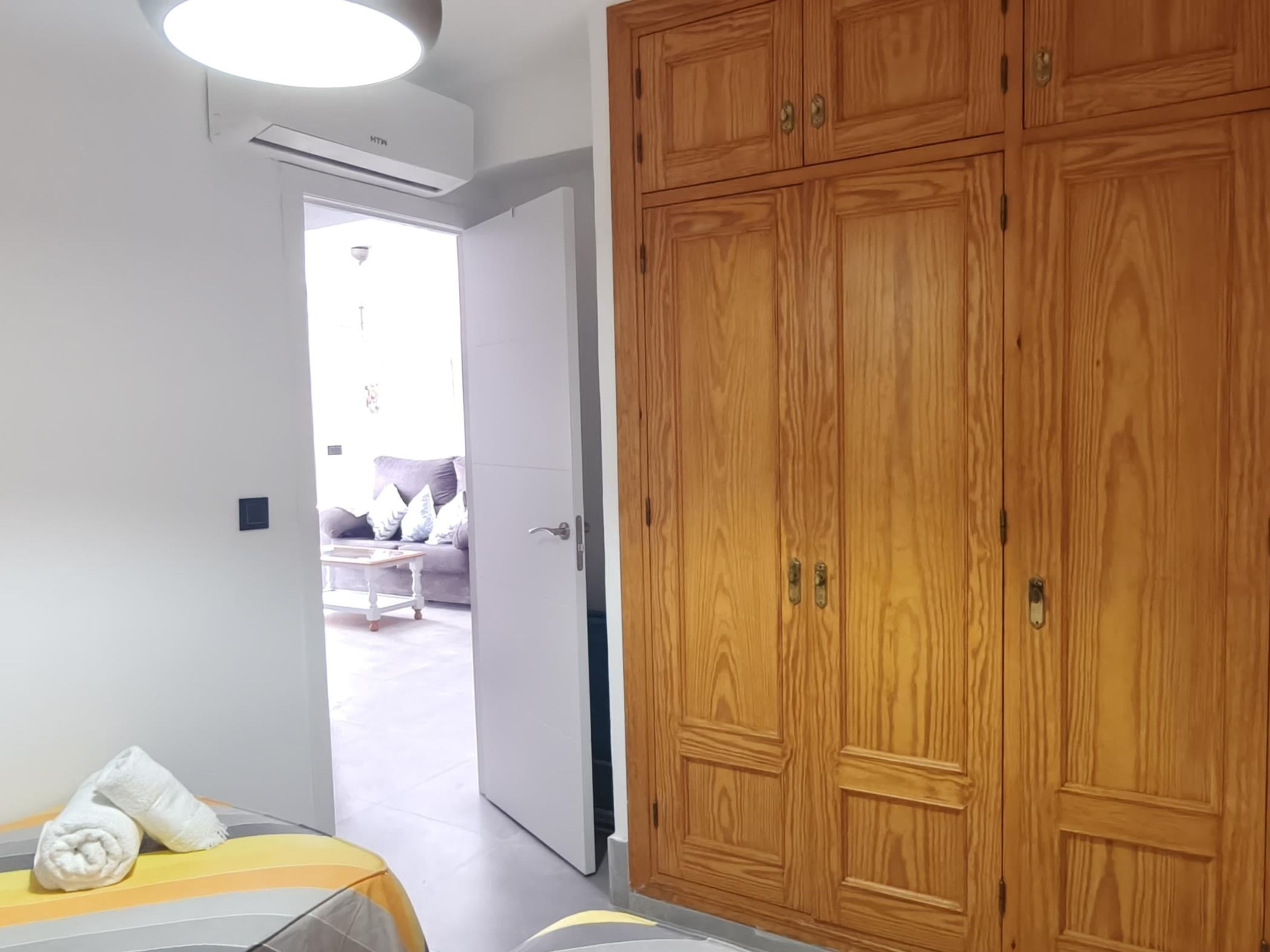 Une chambre d’hôtel avec une armoire en bois, un lit recouvert d’une courtepointe à rayures et une vue sur le salon par une porte ouverte.