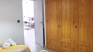 Ein Hotelzimmer mit einem Holzschrank, einem Bett mit gestreifter Bettdecke und Blick in den Wohnbereich durch eine offene Tür.