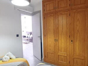 A hotel room with a wooden wardrobe, a bed with a striped bedspread, and a view into the living area through an open door. - Nerja Sun Apartments Bahia 7 (Nerja)