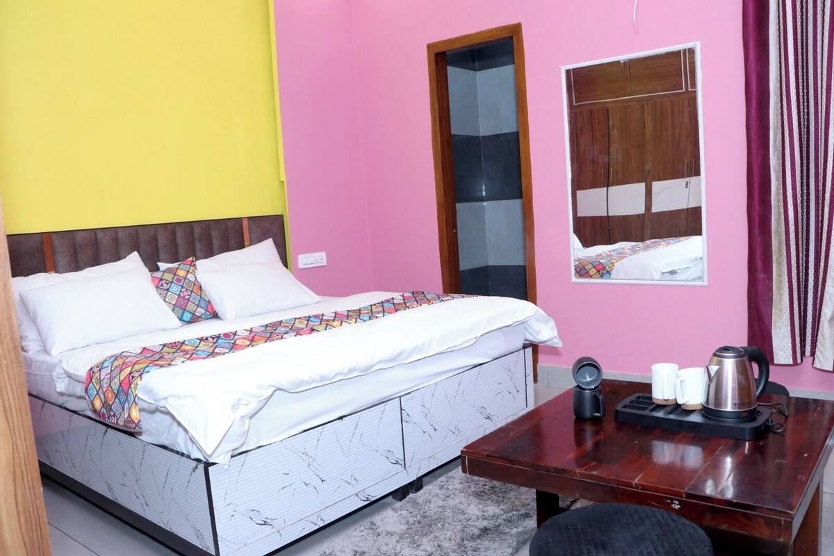 A room with a bed, a wooden table with a kettle and cups, a mirror, and a window with curtains.