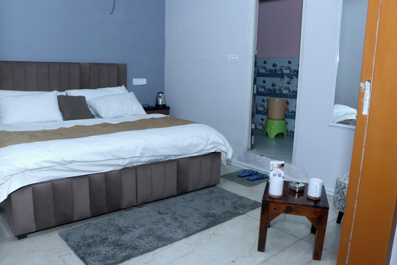 A bedroom with a bed, a nightstand, a coffee table, and a window with a view of a green stool and a patterned wall.