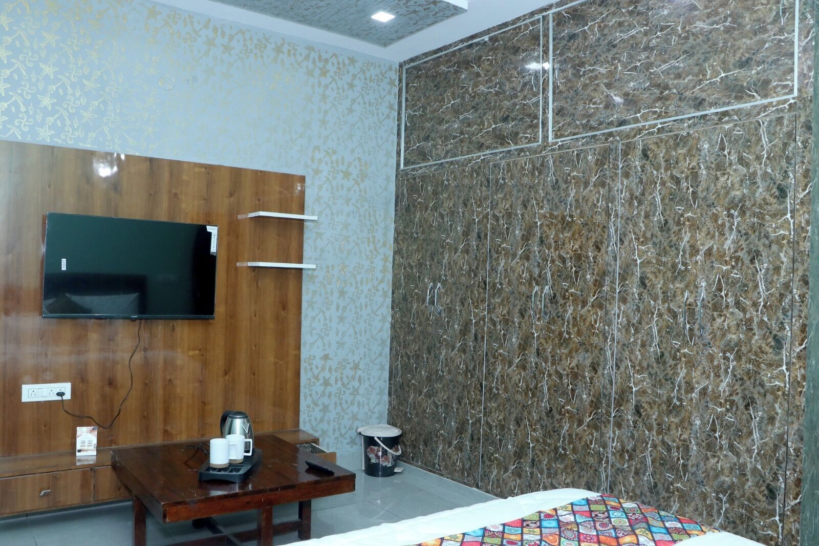 A hotel room with a wooden wardrobe, a flat-screen TV, a marble wall, a bed with a colorful quilt, and a small table with a kettle and cups.