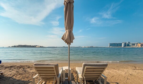 A beach with two lounge chairs and a beach umbrella, overlooking calm waters and distant buildings. - LUX The Grand Bleu Suite (Dubai)