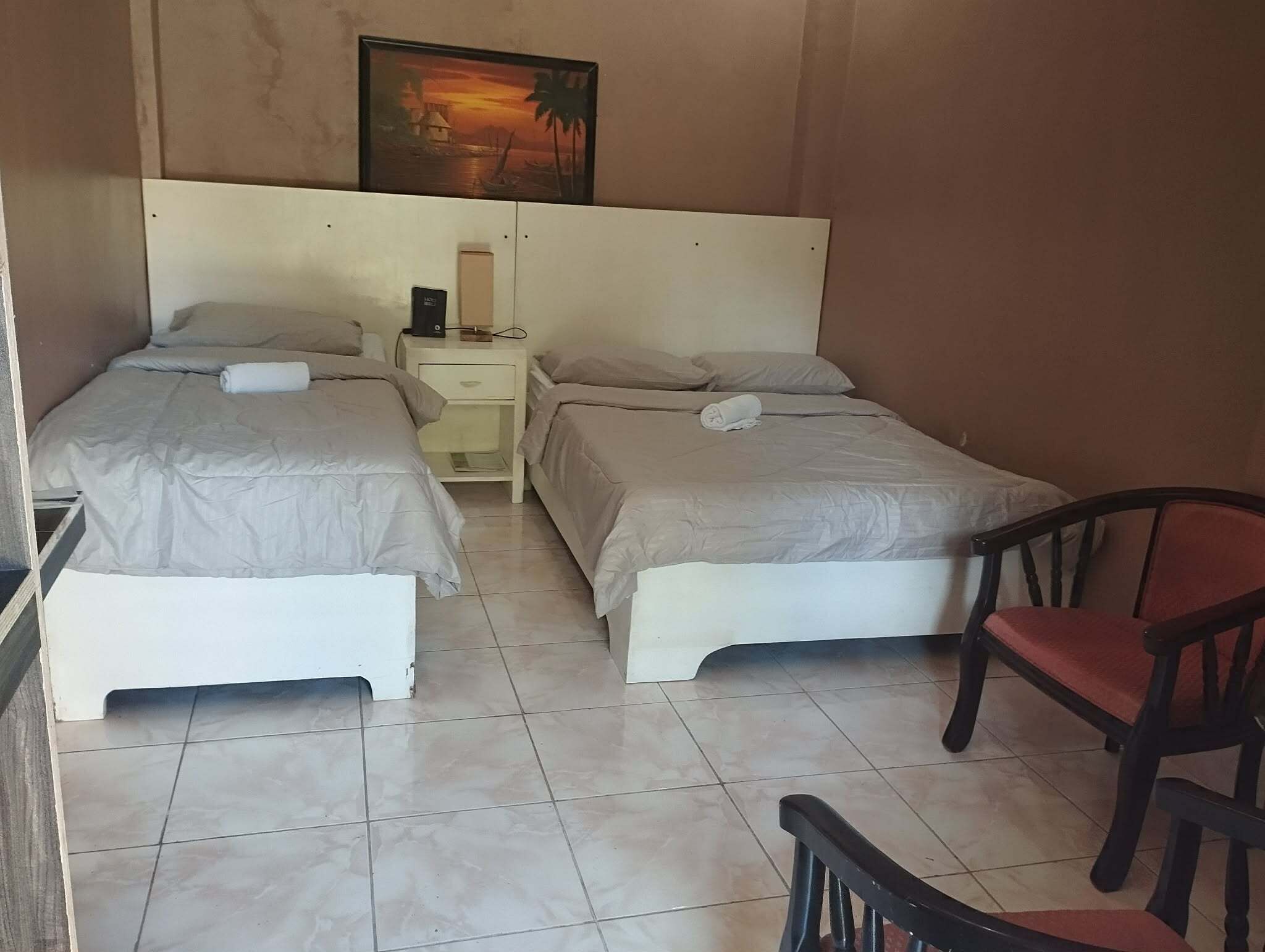 A hotel room with two single beds, a nightstand with a lamp, a chair, and a painting on the wall.