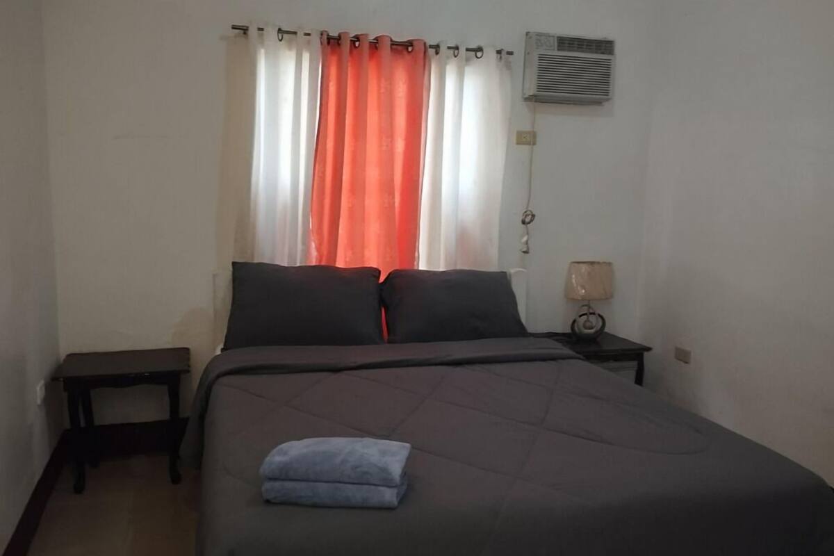 Un lit simple avec une couvre-lit sombre et deux oreillers, une petite table de chevet avec une lampe, et une fenêtre avec des rideaux orange.