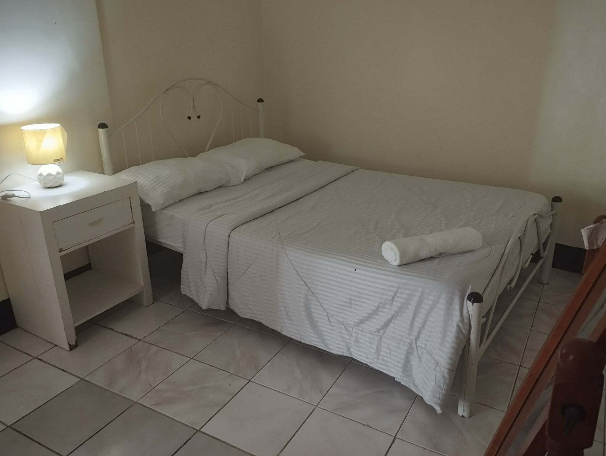 A single-bed room with a white bed, a nightstand with a lamp, and a wooden dresser.