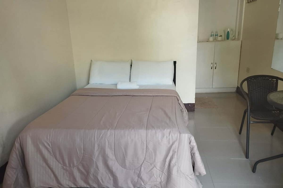 Un lit simple avec une couvre-lit beige, un oreiller blanc et une chaise noire dans une pièce dont le sol est carrelé et qui contient une armoire avec des articles de toilette.