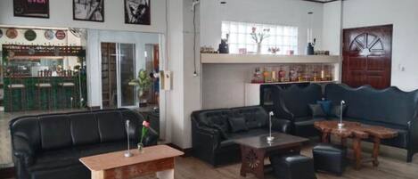 Amplia sala de estar con un sofá de cuero negro, una mesa de centro de madera y un sofá de cuero oscuro. Se observa una mesa de comedor de madera con sillas, y un área de bar visible a través de las puertas de cristal.