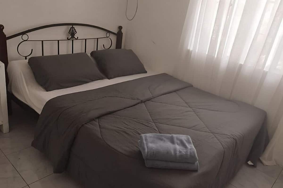 Un lit simple avec une couette marron foncé et deux oreillers, un mur blanc et une fenêtre avec des rideaux légers.