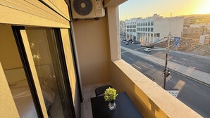 Een balkon met een tafel en stoelen, een airconditioning en uitzicht op een stadsweg tijdens zonsondergang.