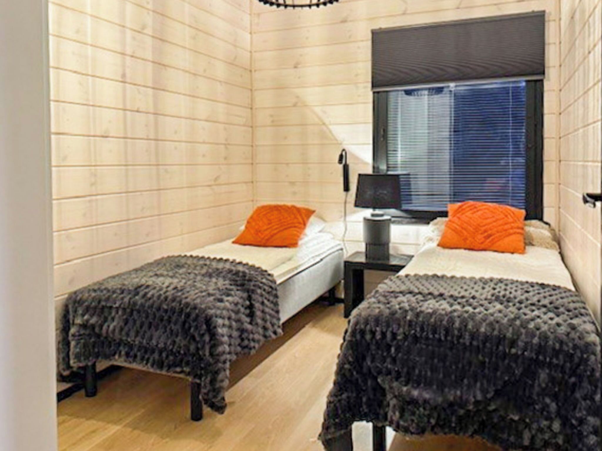 Une petite pièce agréable, dotée de deux lits, chacun agrémenté d’un oreiller orange et d’une couverture grise. On y trouve une fenêtre avec des stores, un bureau avec une lampe et une chaise.