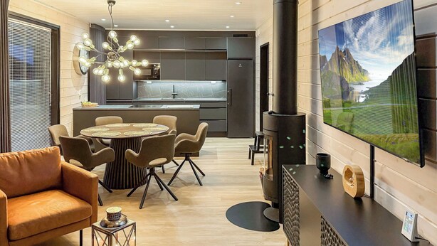 A modern kitchen with a central island, a dining area with a round table and chairs, a large flat-screen TV mounted on the wall, and a wood-burning stove. - Saanan taika by Interhome (Enontekiö)